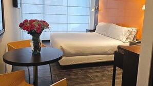 A hotel room with a bed, a small table with a vase of flowers, a chair, and a window with blinds. - 104 Art Suites (Bogotá)