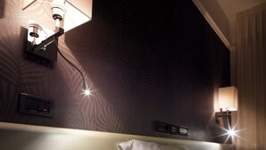 A hotel room with a bed, a bedside table with an open book, and wall-mounted lamps.