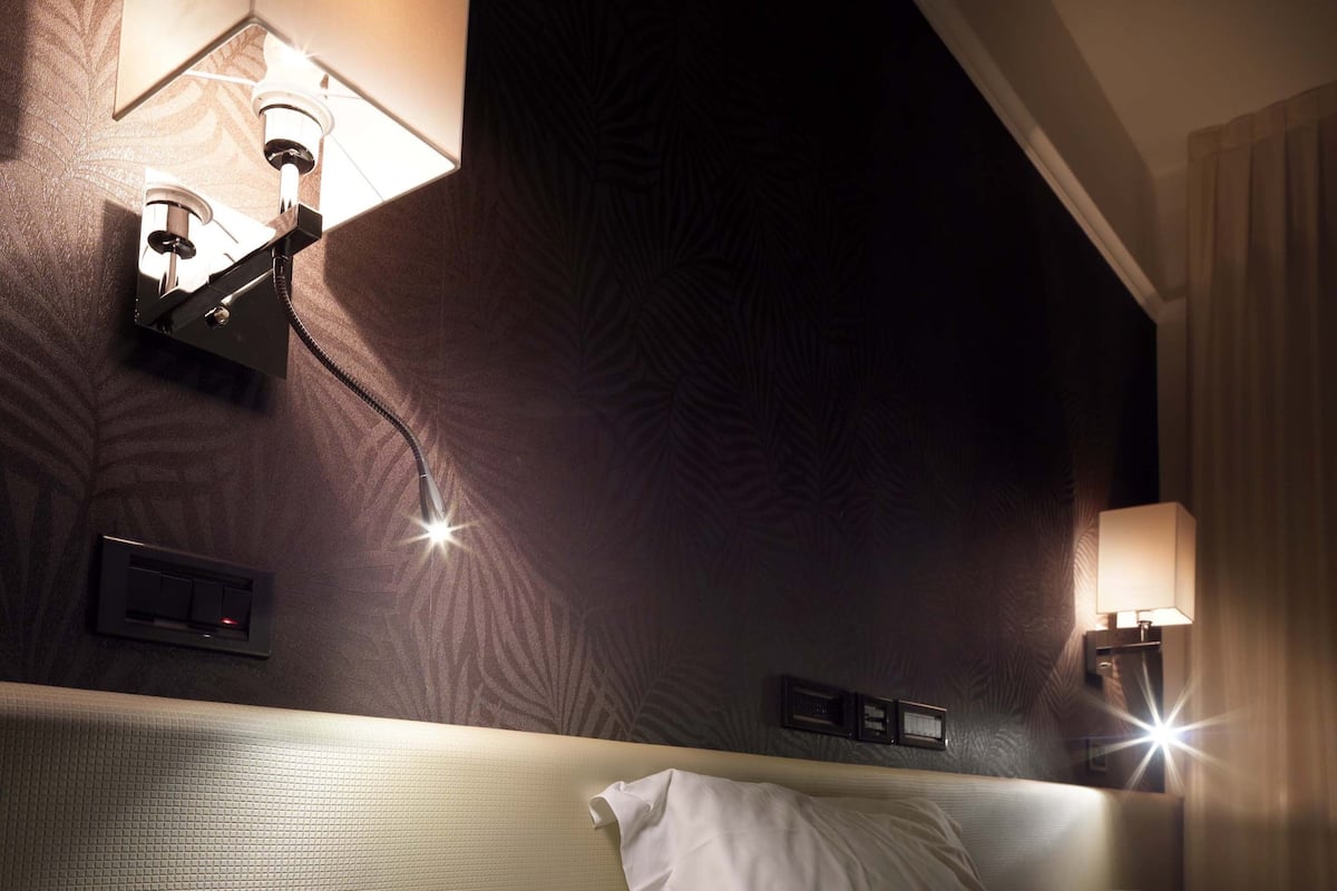 A hotel room with a bed, a bedside table with an open book, and wall-mounted lamps.