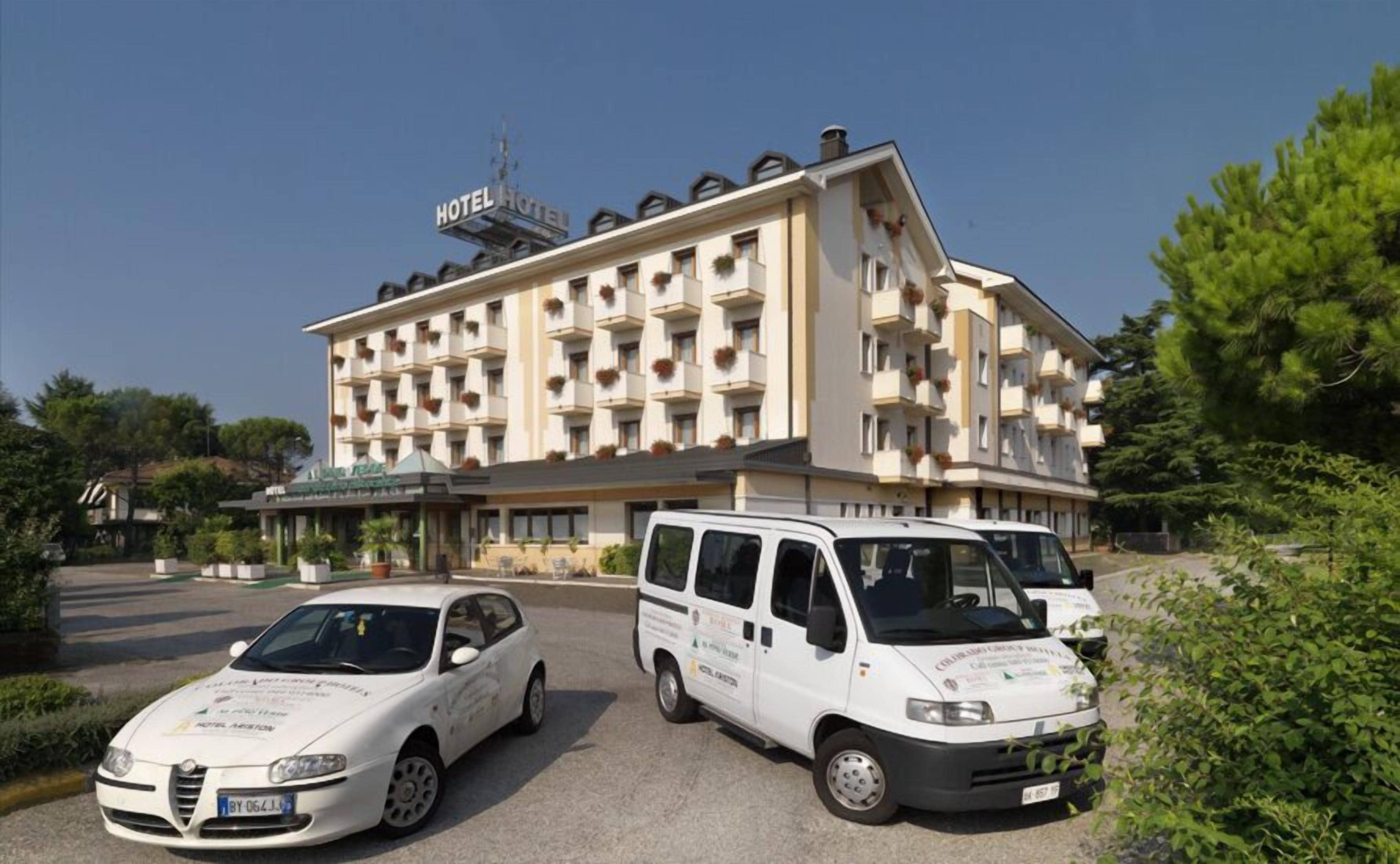 A hotel with a modern facade, multiple balconies, and two white service vehicles parked in front.