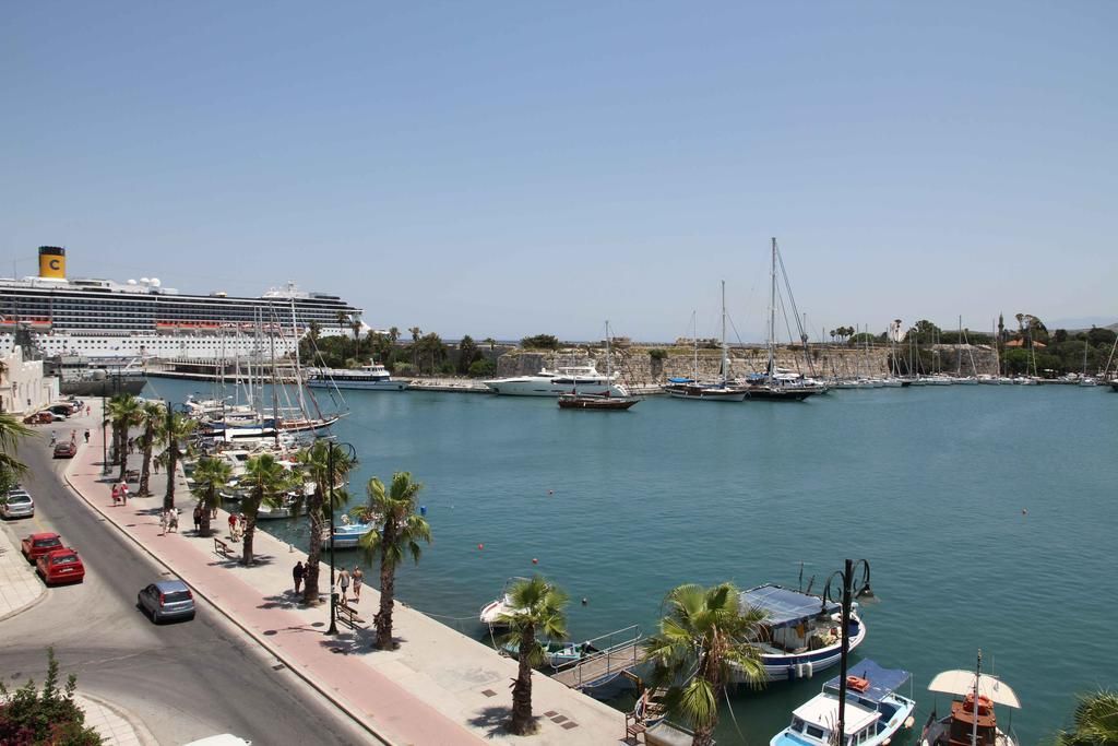 Un crucero amarrado en una marina con varios barcos y yates, un paseo marítimo pavimentado con palmeras y un cielo despejado.