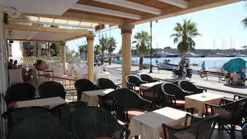 An outdoor cafe with tables and chairs, overlooking a marina with boats and palm trees.