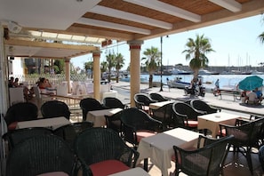 Un café al aire libre con mesas y sillas, con vistas a un puerto deportivo con barcos y palmeras.