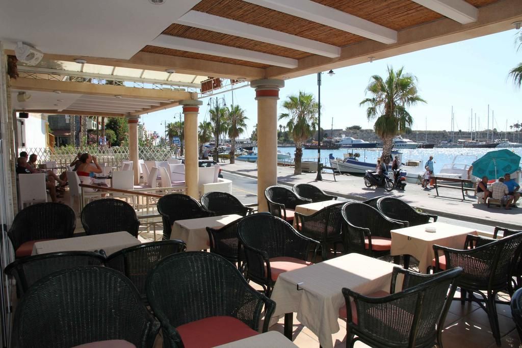 An outdoor cafe with tables and chairs, overlooking a marina with boats and palm trees.