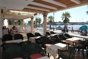 An outdoor cafe with tables and chairs, overlooking a marina with boats and palm trees.