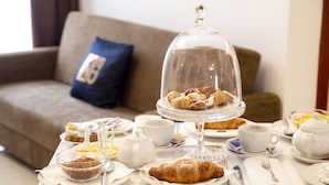 Une table dressée pour le petit-déjeuner, avec des viennoiseries, du thé et du café, dans une pièce où se trouvent un canapé et un coussin bleu.