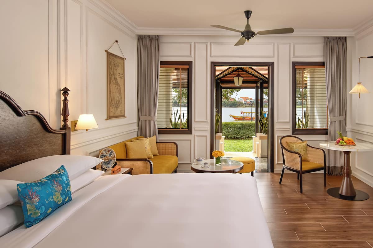A hotel room with a bed, a sofa, a dining table, and a view of a garden.