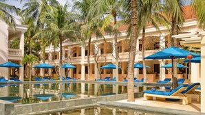 A pool area with lounge chairs and umbrellas, surrounded by palm trees and a building with balconies.