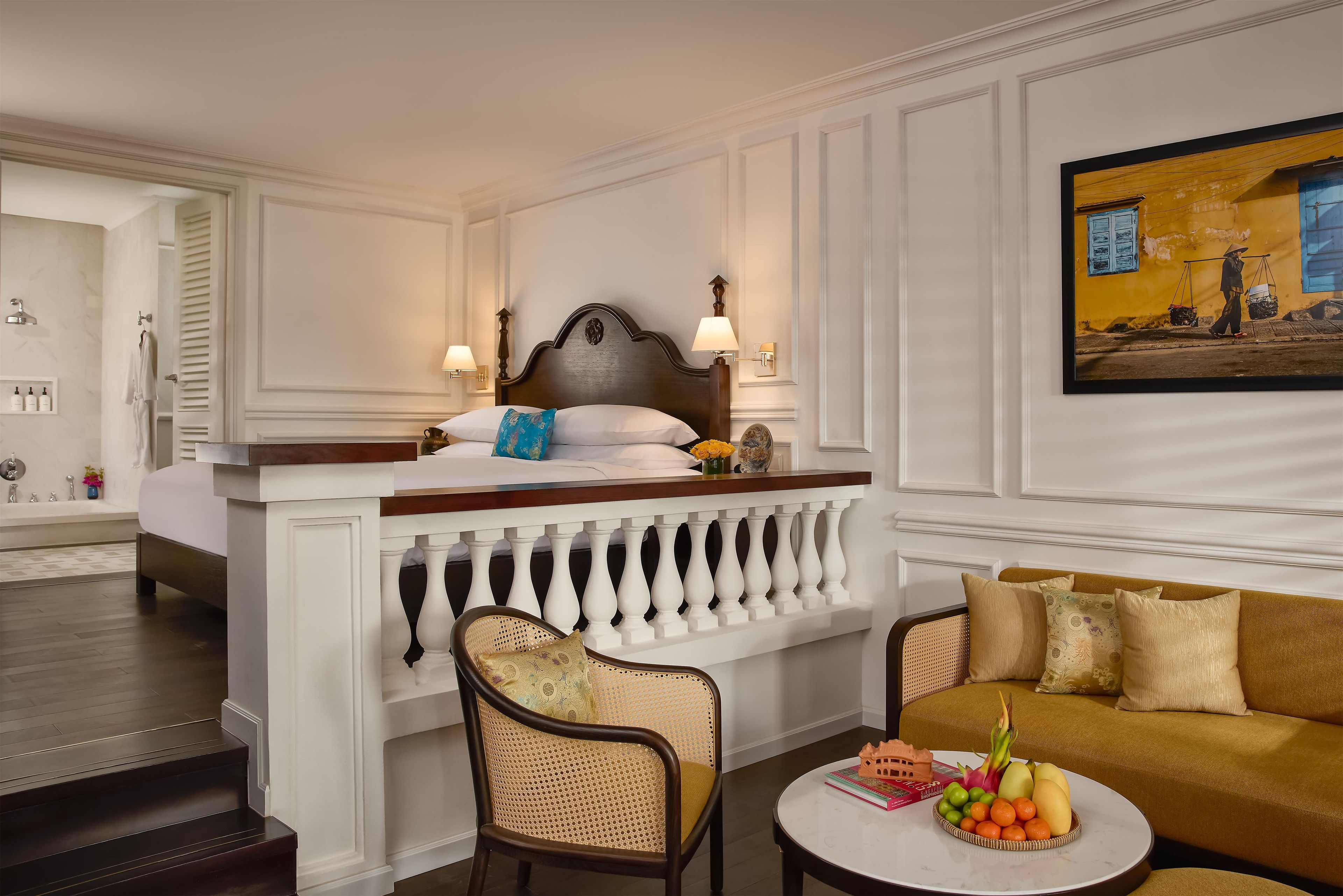 A hotel room with a bed, a sitting area with a sofa and a chair, a small table with fruit and books, and a painting on the wall.