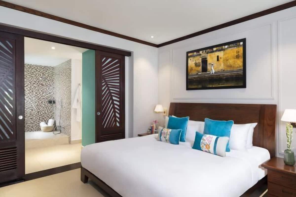 A hotel room with a large bed, a wooden headboard, a painting on the wall, and a bathroom visible through an open door.
