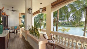 A wooden deck with a railing, a seating area with chairs, a table, and a view of a body of water with palm trees. - Anantara Hoi An Resort (Da Nang)
