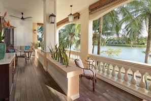 A wooden deck with a railing, a seating area with chairs, a table, and a view of a body of water with palm trees. - Anantara Hoi An Resort (Da Nang)