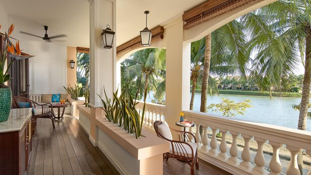 A wooden deck with a railing, a seating area with chairs, a table, and a view of a body of water with palm trees.