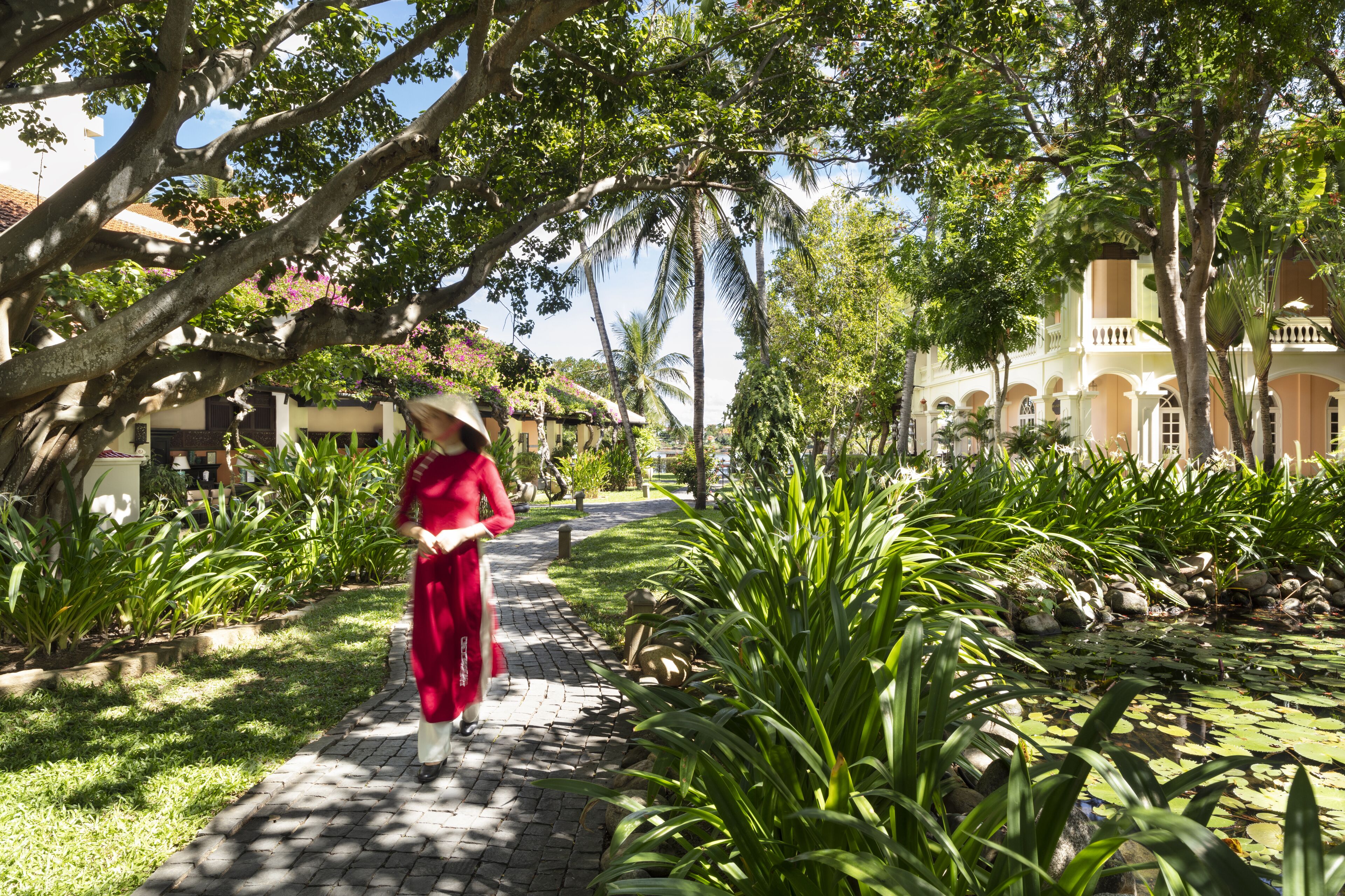 Un camino bordeado de exuberante vegetación y palmeras, que conduce a un edificio de estilo arquitectónico tradicional.
