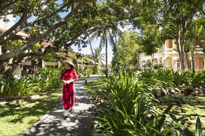 Uma trilha bordeada por vegetação exuberante e palmeiras, que conduz a um edifício com arquitetura tradicional.