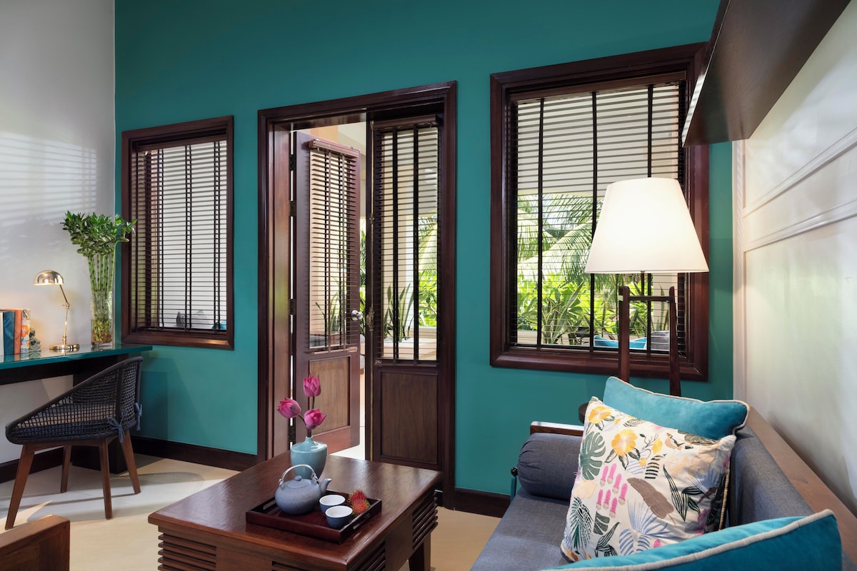 A room with teal walls, wooden shutters, a sofa with a floral pillow, a table with a teapot, and a chair.