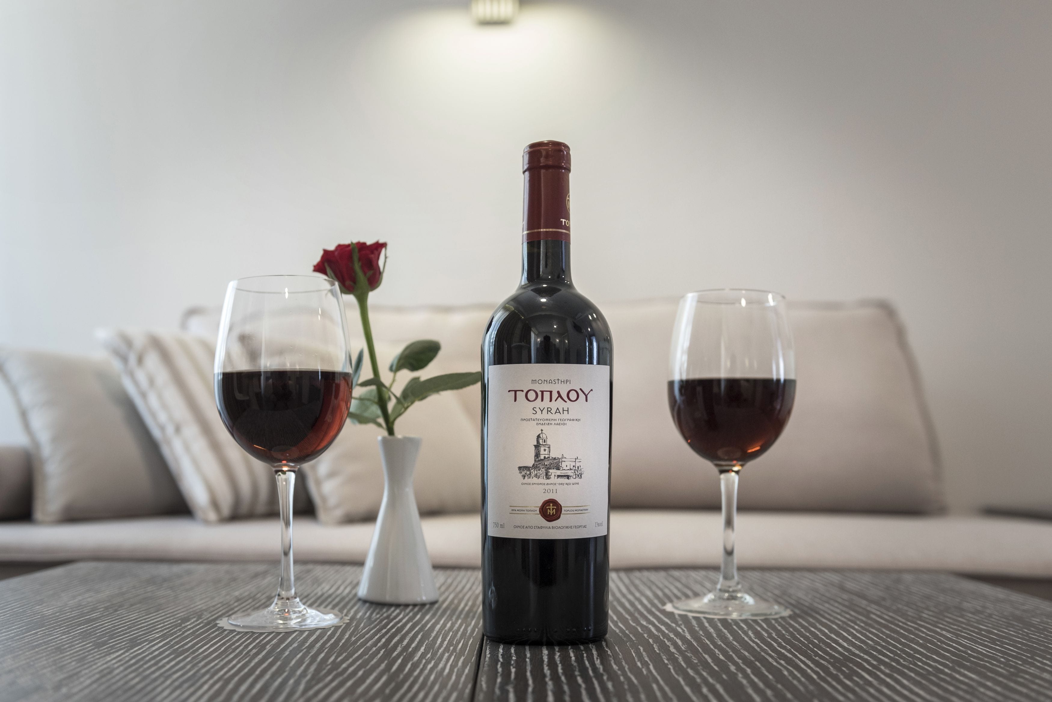 A wine bottle and two glasses on a table with a rose and a vase in the background.