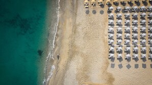 Vista aérea de una playa con hileras de sombrillas y tumbonas, una orilla arenosa y un mar azul transparente.