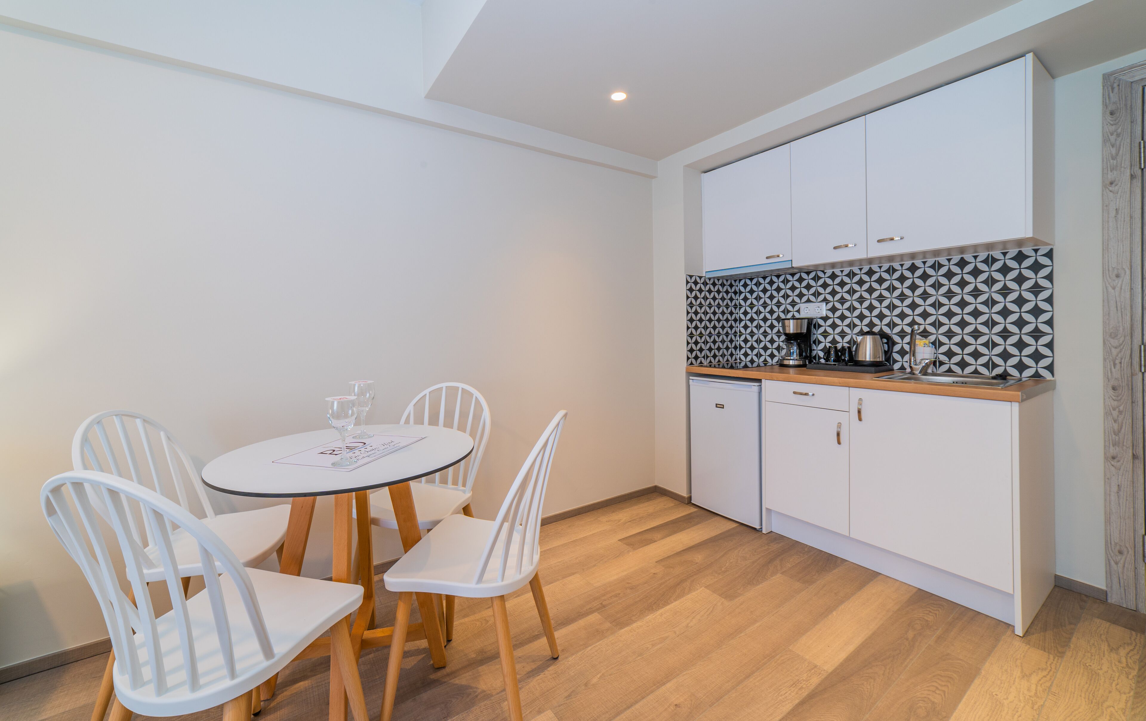 Uma cozinha compacta com armários brancos, backsplash estampado e um pequeno espaço de jantar com mesa redonda e cadeiras brancas.