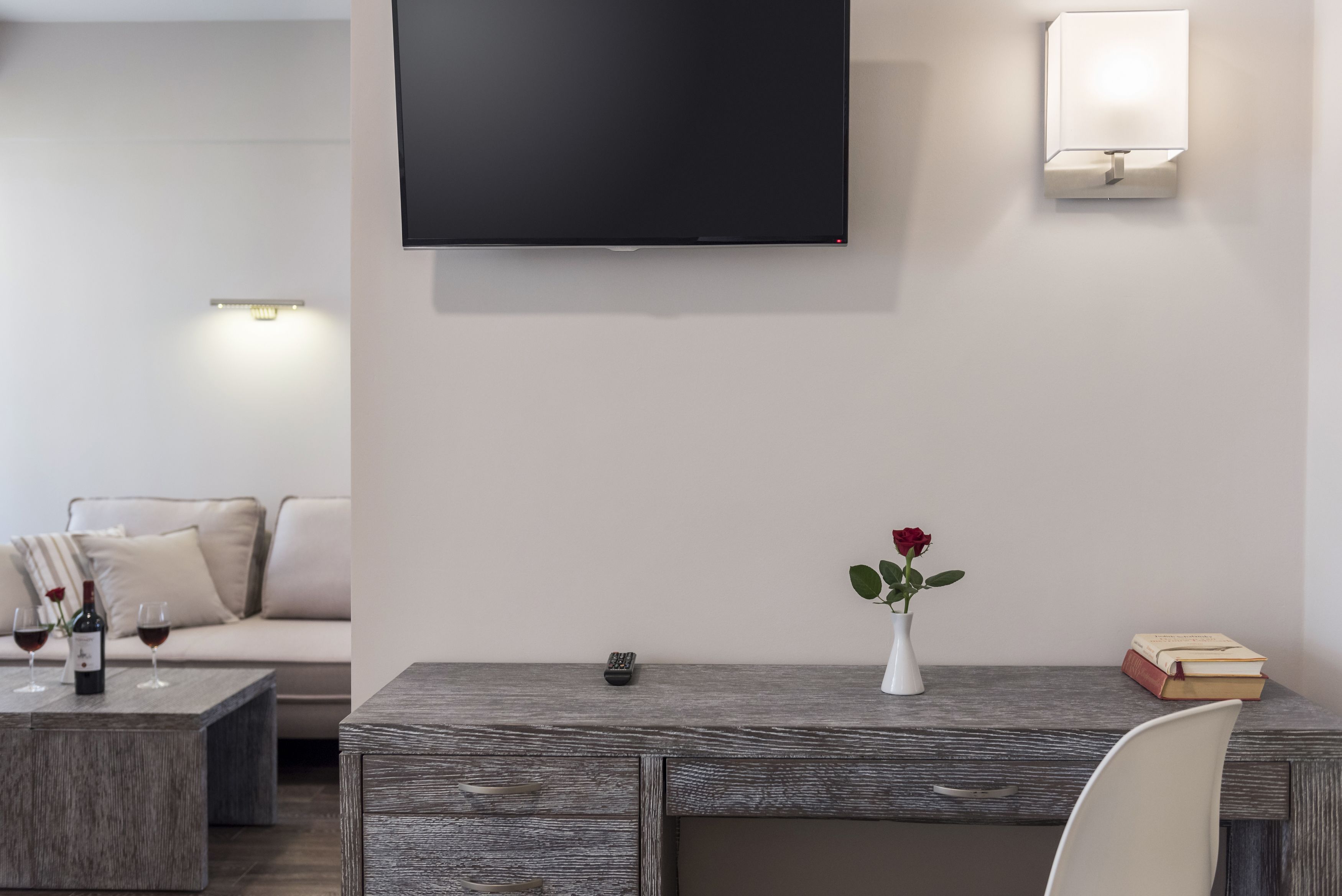 A modern living room with a flat-screen TV mounted on the wall, a wooden coffee table with wine glasses and a remote control, a white vase with a rose, and a bookshelf with books and a lamp.