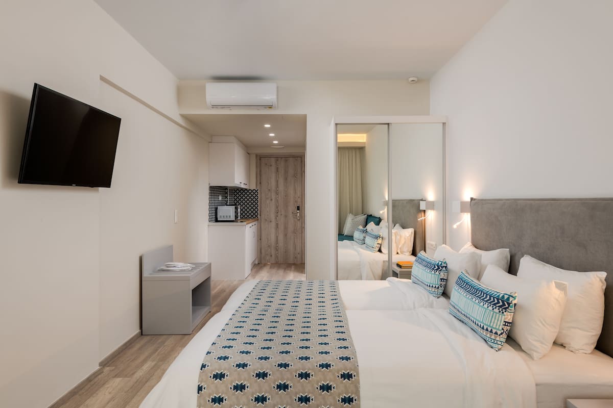 Et moderne hotelværelse med en stor seng, et fladskærms-tv monteret på væggen, et lille tekøkken og et spejl der reflekterer rummet.