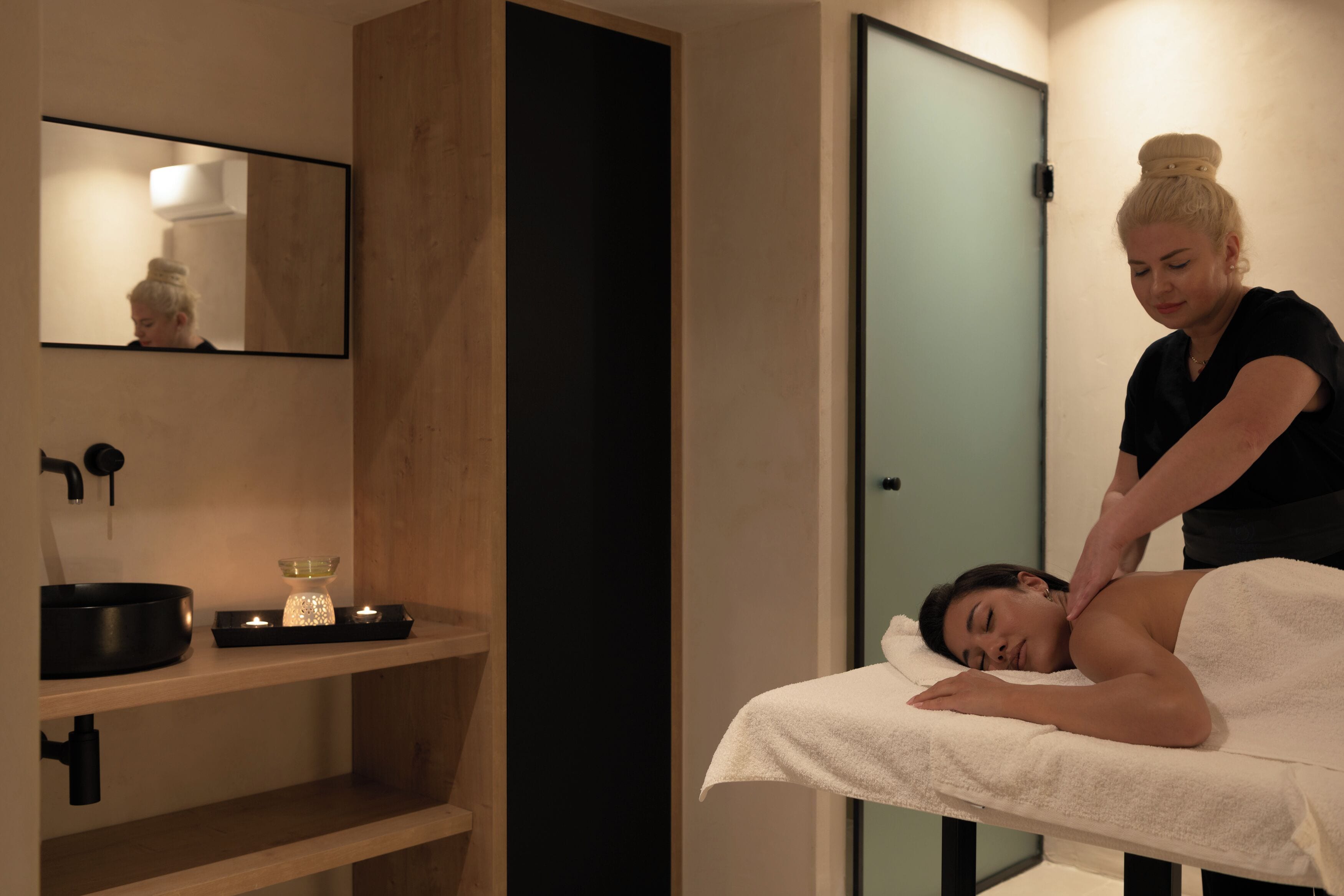 A spa room with a massage table, a mirror, a sink, and a shelf with candles.