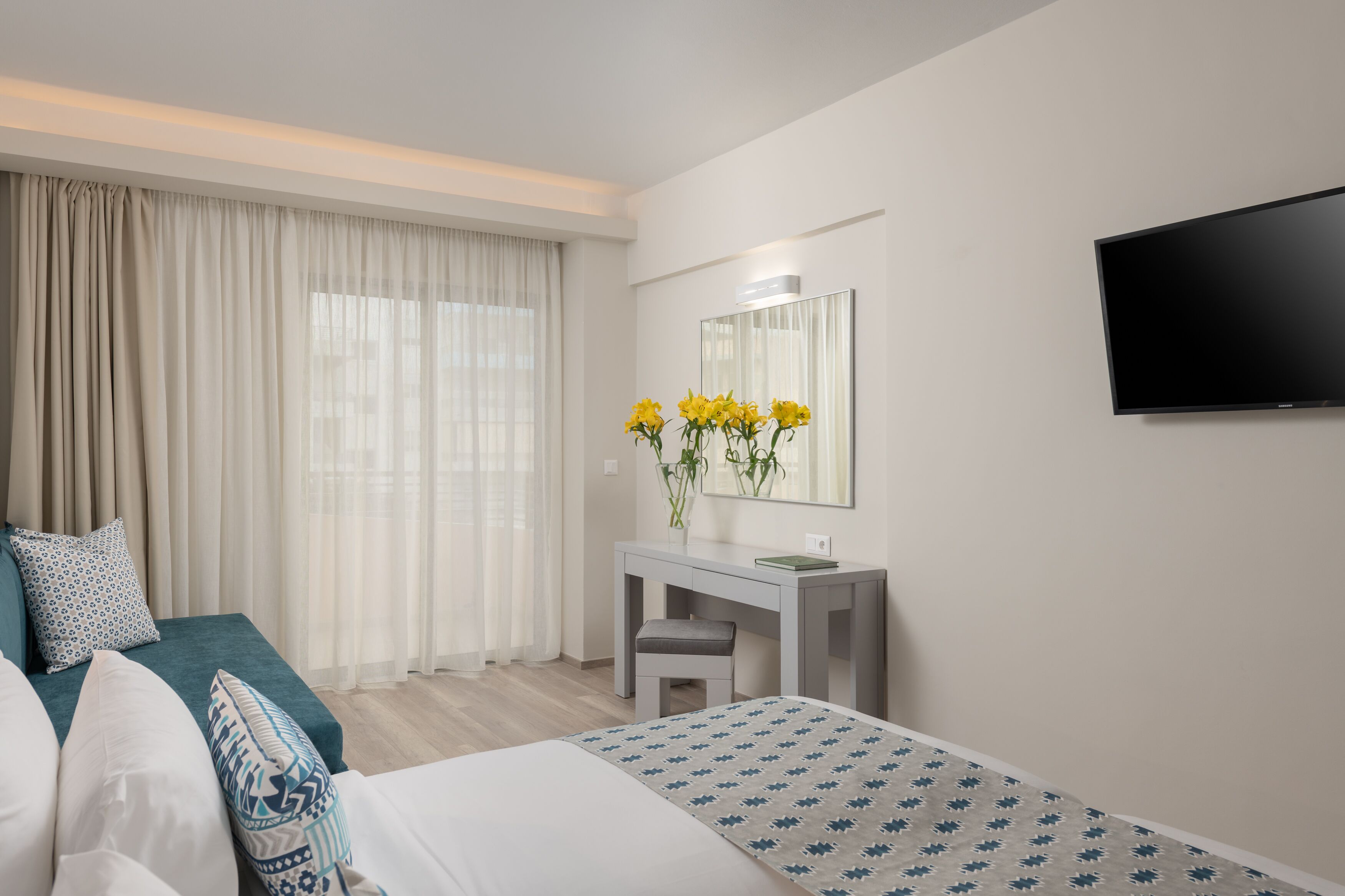 A modern hotel room with a bed, a desk with a mirror, a TV, and a window with curtains.