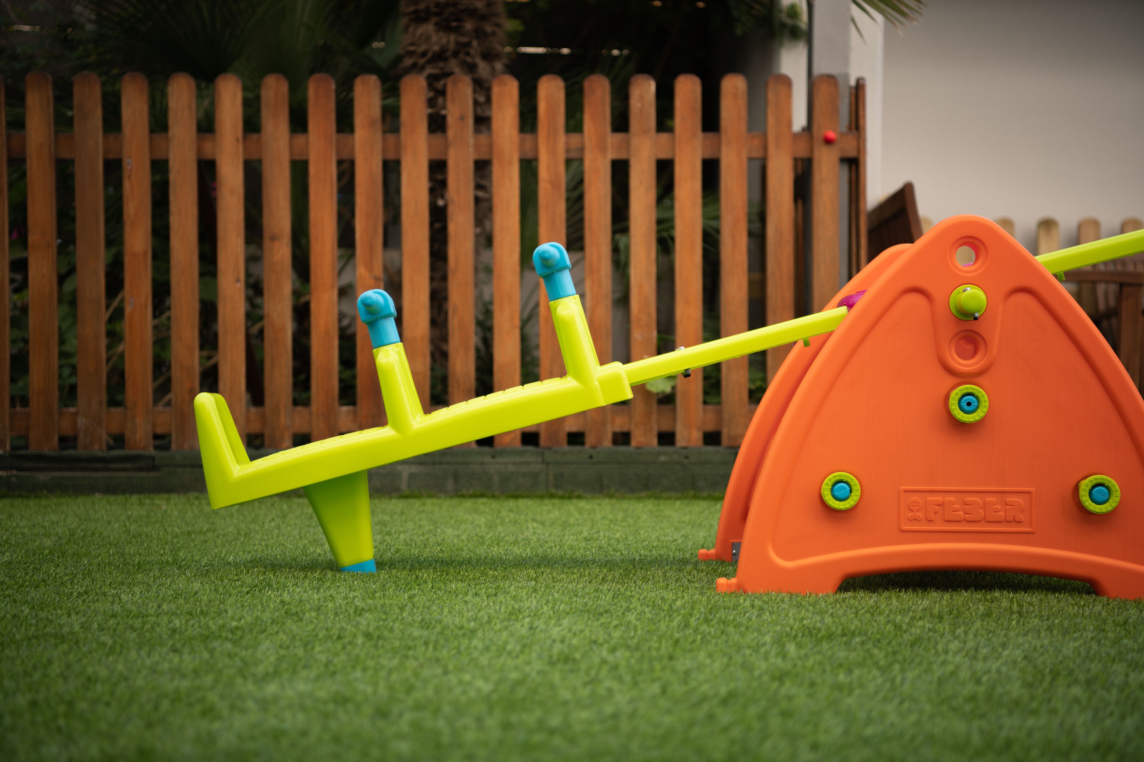 A seesaw on artificial grass with a wooden fence in the background.
