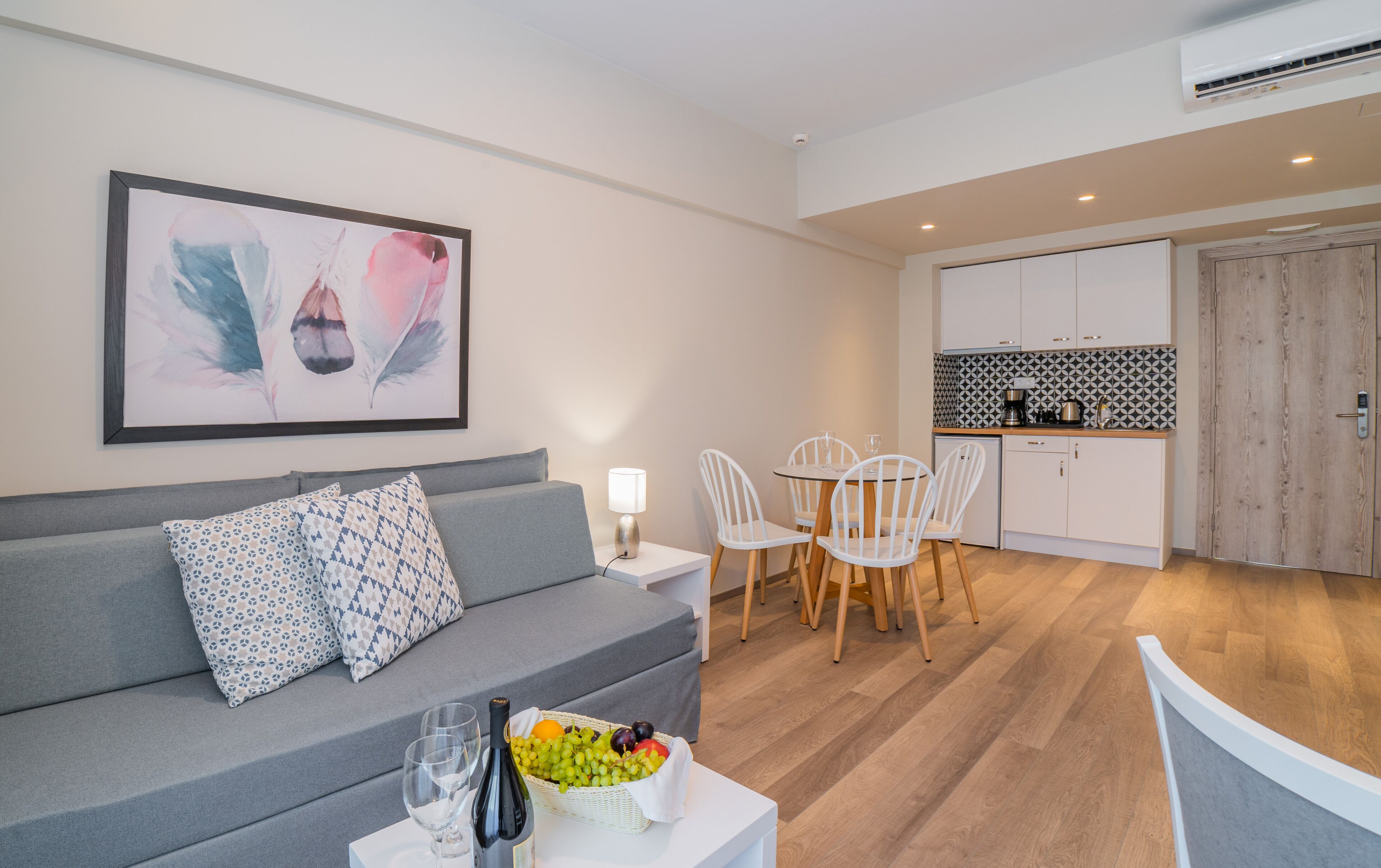 A modern living room with a grey sofa, a dining table with white chairs, and a kitchen area with white cabinets and a patterned backsplash.