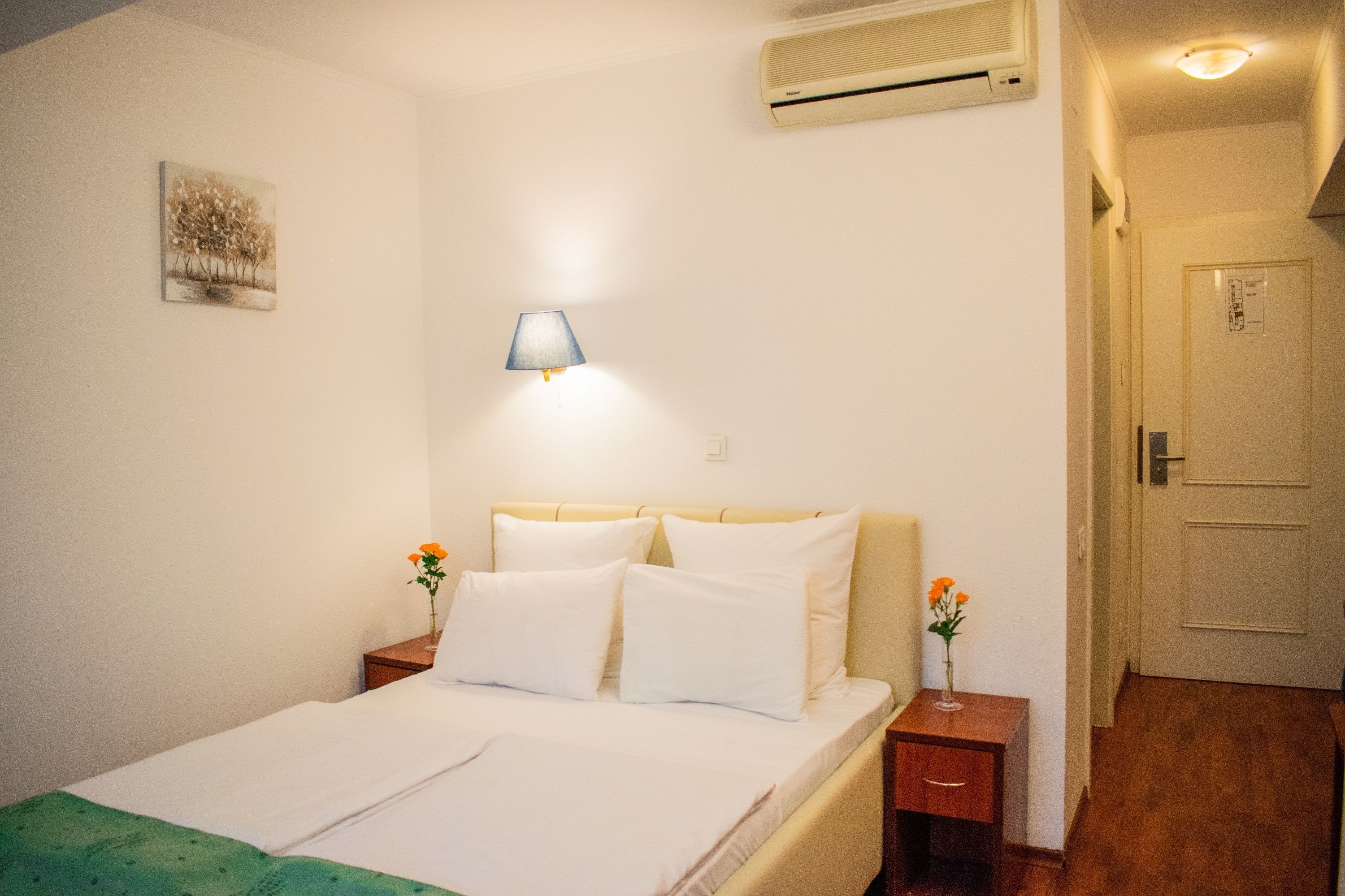 A hotel room with a bed, bedside tables, a lamp, an air conditioner, and a door.