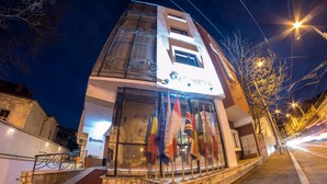 Un edificio moderno con fachada de vidrio, alfombra roja en la entrada y varias banderas a la vista.