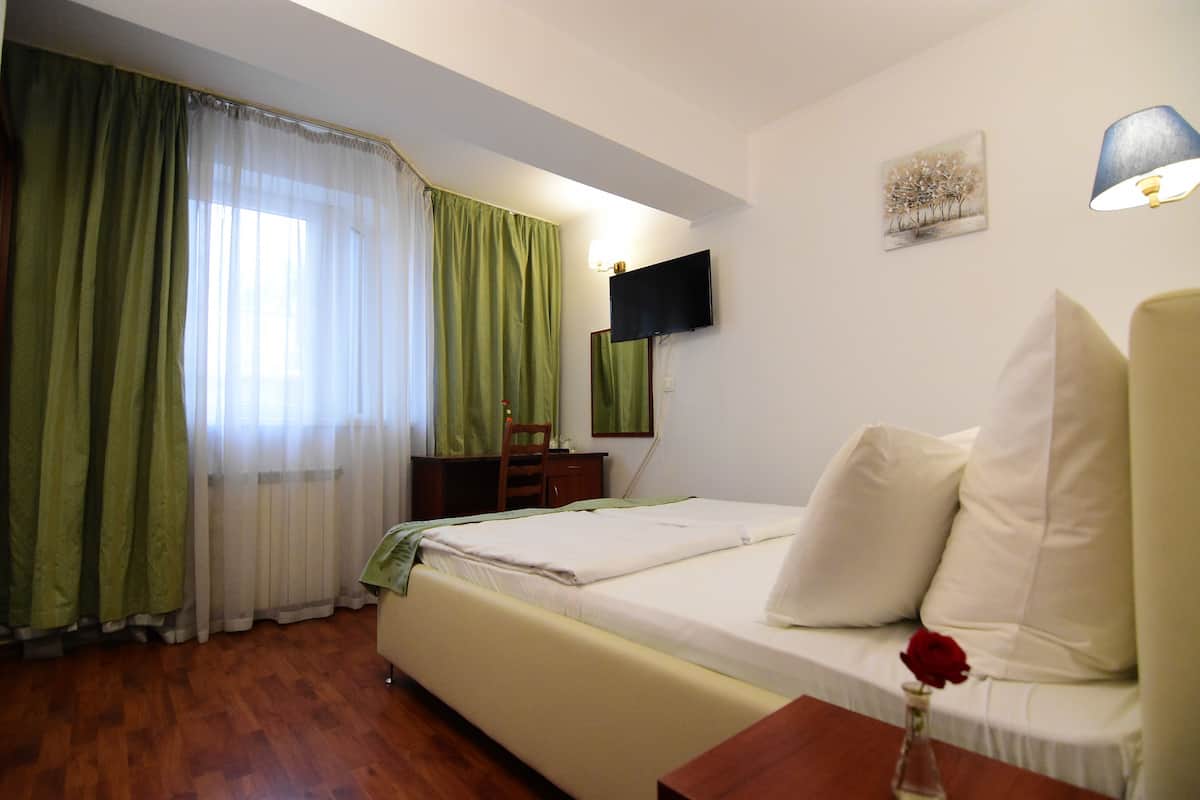 A hotel room with a bed, a desk, a chair, a TV, and a vase with a flower.