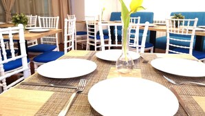 Un área de comedor con sillas blancas, cojines azules y una mesa central con un florero transparente y un plato.