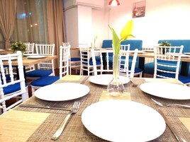 Un área de comedor con sillas blancas, cojines azules y una mesa central con un florero transparente y un plato.