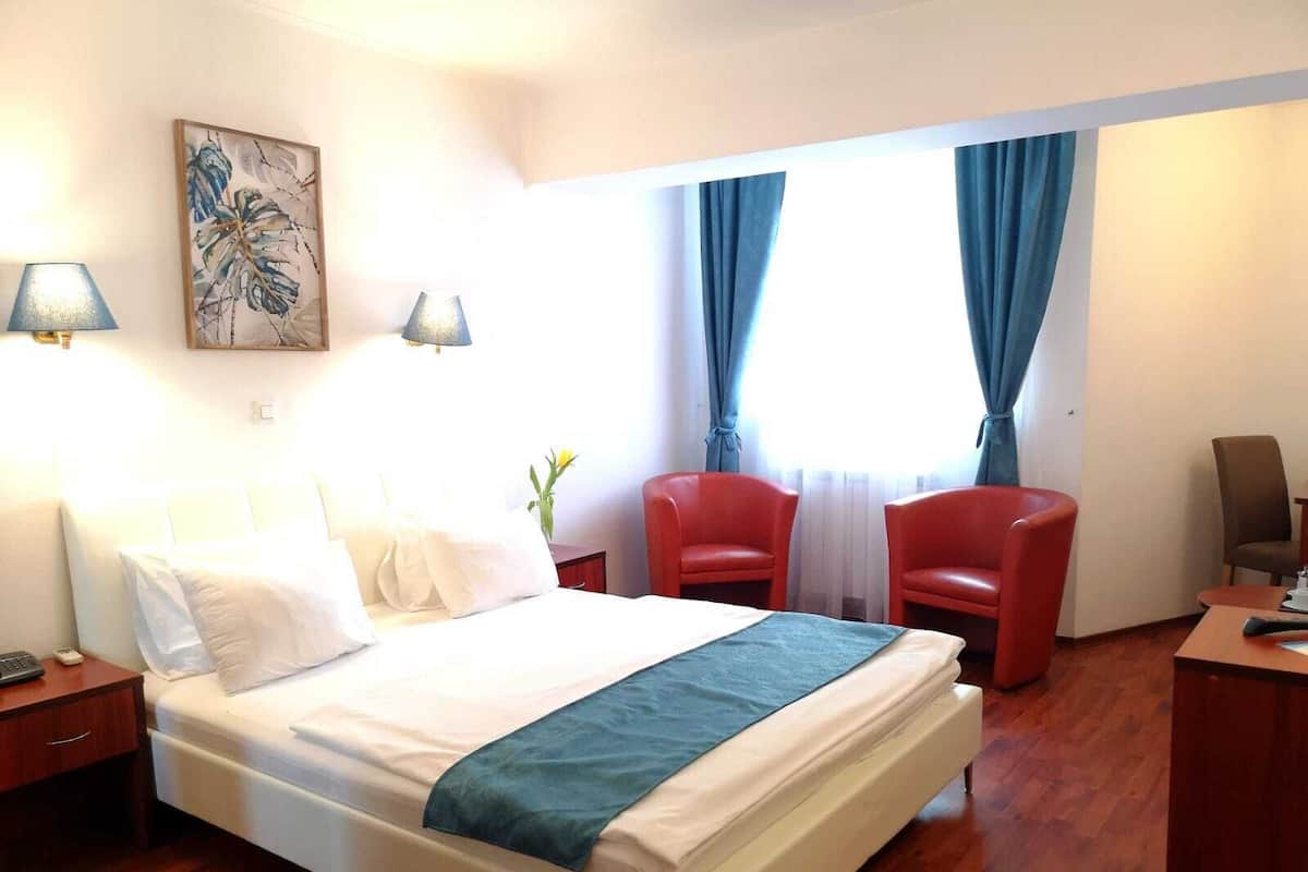A hotel room with a bed, two red chairs, a desk with a lamp, and a painting on the wall.