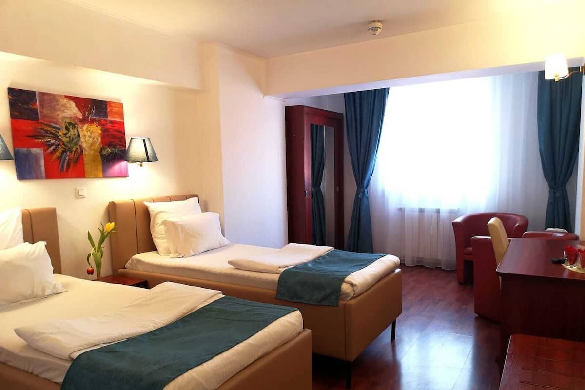 A hotel room with two beds, a desk, a chair, a painting, and a window with curtains.