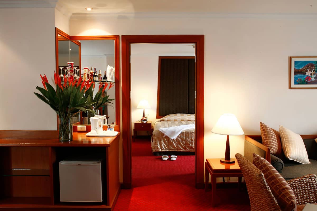 A hotel room with a bed, a desk with a mirror, a small fridge, a bedside table with a lamp, a chair, and a decorative plant.