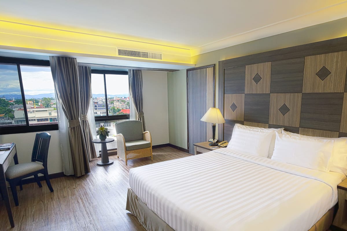 A hotel room with a large bed, a desk with a chair, a small table with a lamp, and a view of the city through large windows.