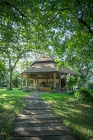 Um gazebo de madeira com telhado de palha, rodeado por vegetação exuberante e um caminho de pedra que leva até ele.