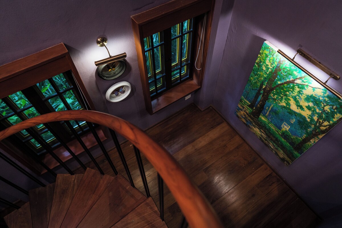 Una escalera de madera con barandilla, una ventana que da a zonas verdes y un cuadro de un bosque en la pared.