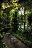Um pátio com mesa e cadeiras, cercado por vegetação exuberante e um edifício com várias janelas.