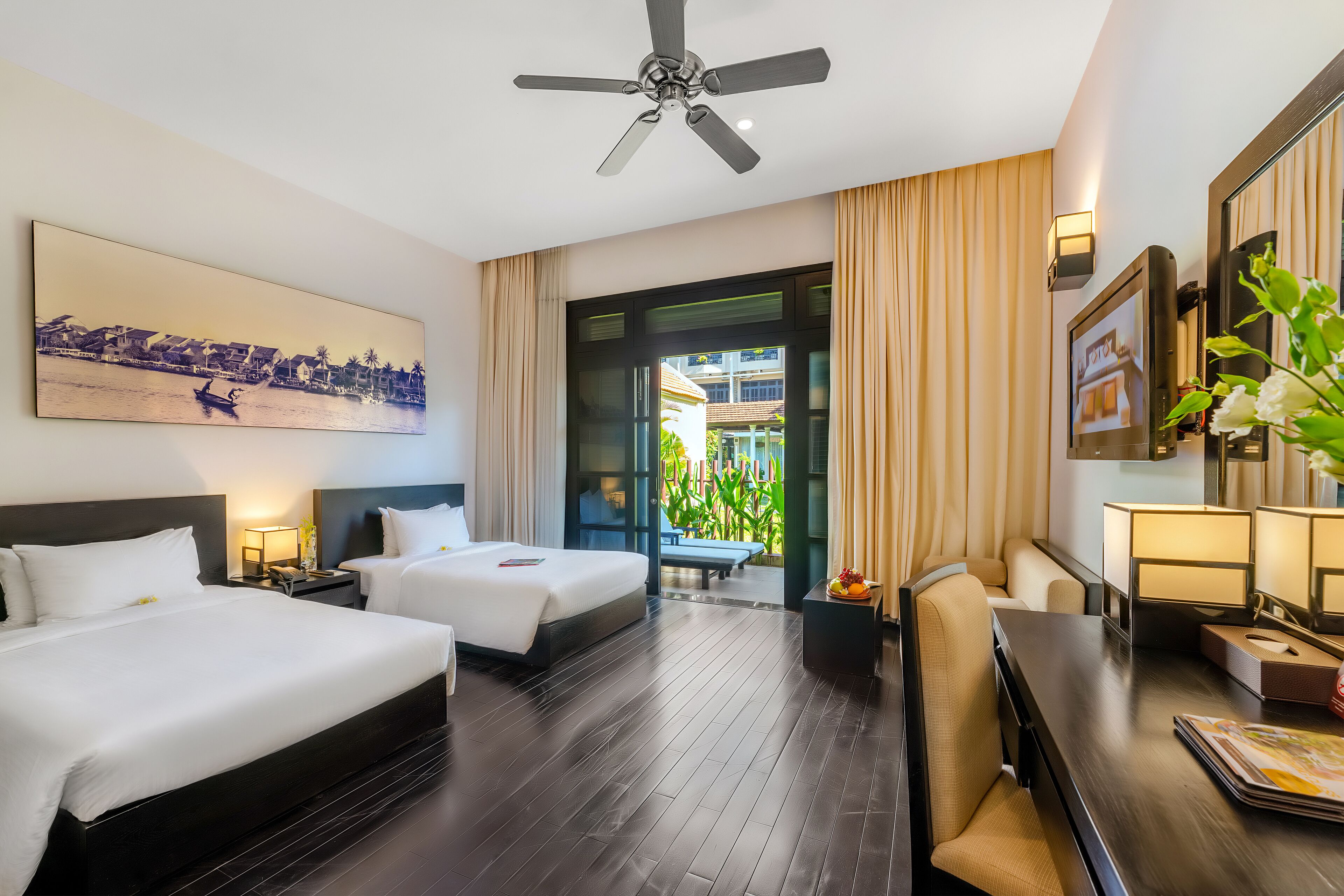 Habitación de hotel con dos camas, un escritorio, una silla, un ventilador de techo, un ventanal y vistas a un paisaje urbano.