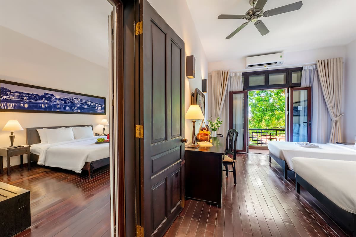 A hotel room with two beds, a desk with a lamp, a cityscape painting, and a balcony with a view of greenery.
