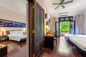 A hotel room with two beds, a desk with a lamp, a cityscape painting, and a balcony with a view of greenery. - Hoi An Historic Hotel (Da Nang)