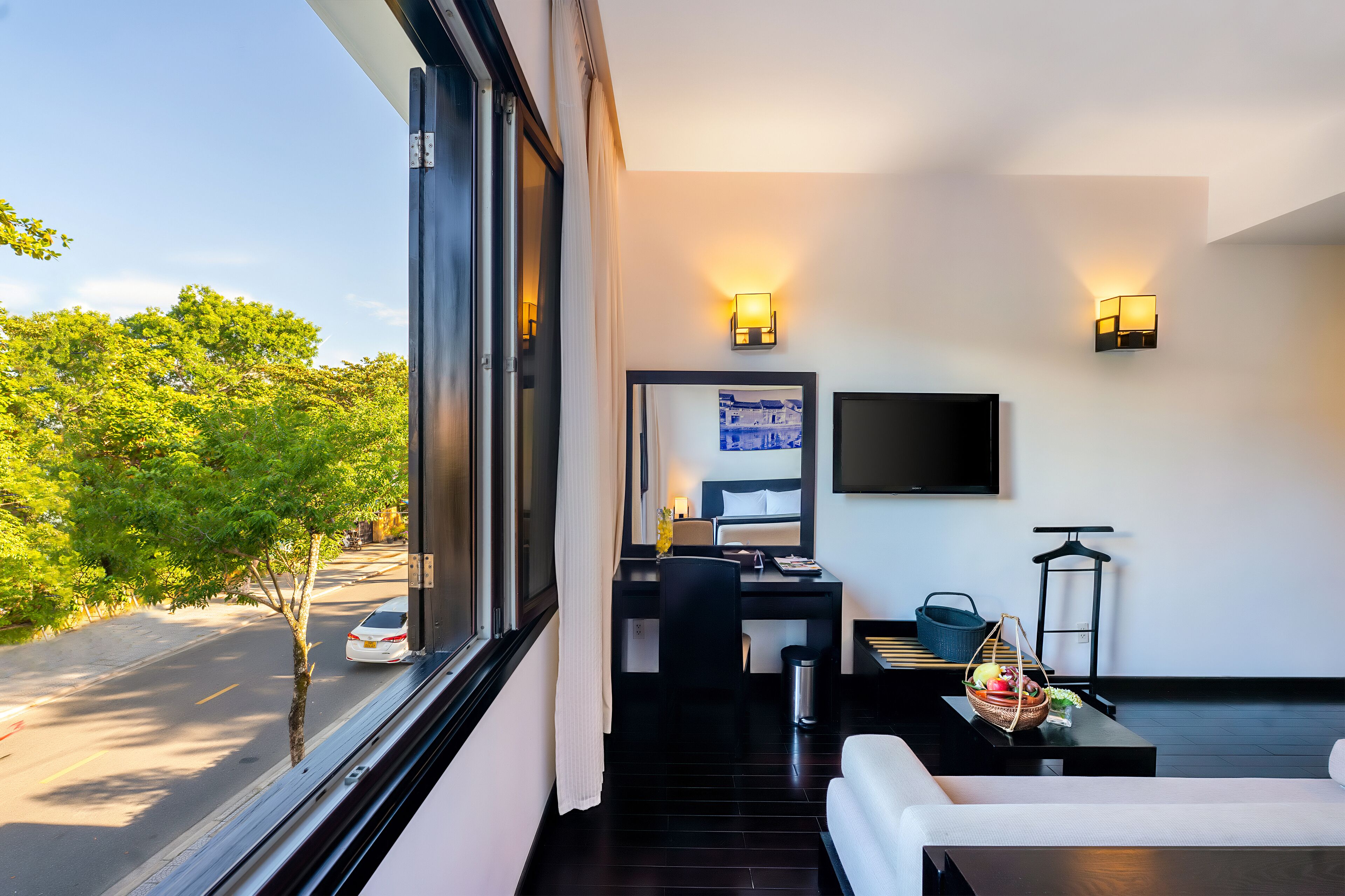 A modern hotel room with a large window, a desk with a chair, a sofa, and a flat-screen TV mounted on the wall.