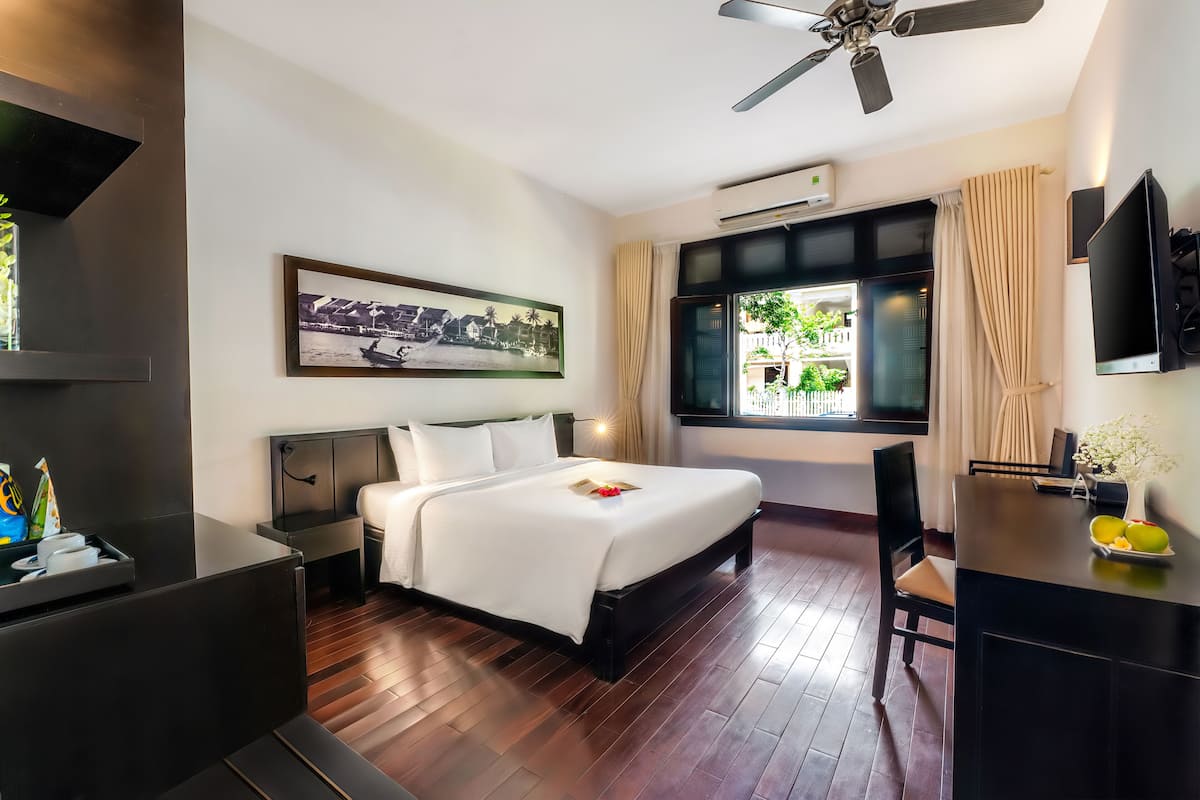 A hotel room with a bed, a desk, a chair, a ceiling fan, a window with curtains, a framed picture on the wall, and a TV on the wall.