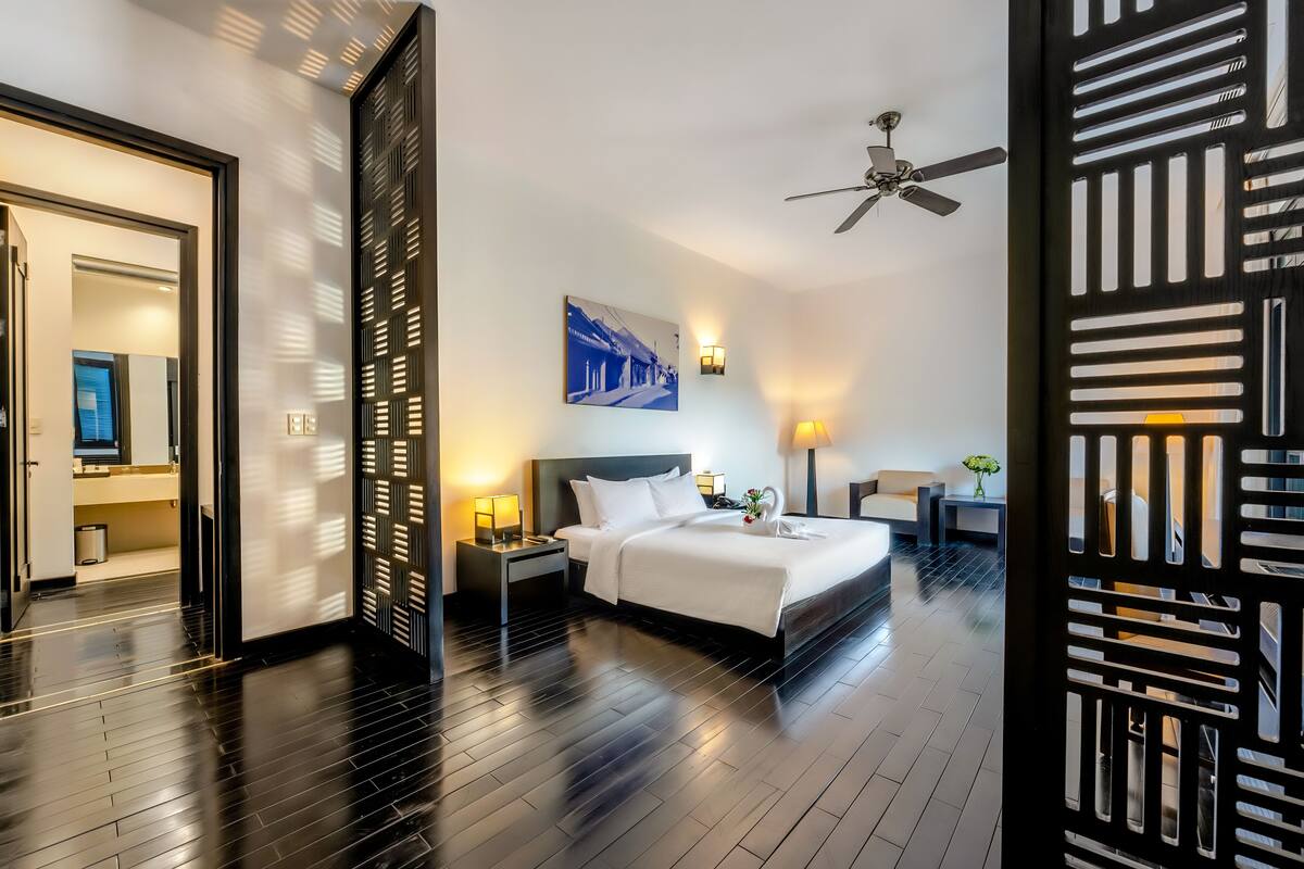 A modern hotel room with a large bed, a ceiling fan, a nightstand with a lamp, and a wall-mounted picture.