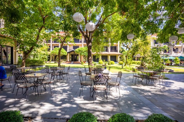 An outdoor patio area with tables and chairs, surrounded by trees and buildings. - Hoi An Historic Hotel (Da Nang)