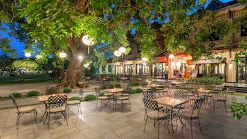 An outdoor cafe area with tables and chairs, surrounded by trees and illuminated by hanging lights.