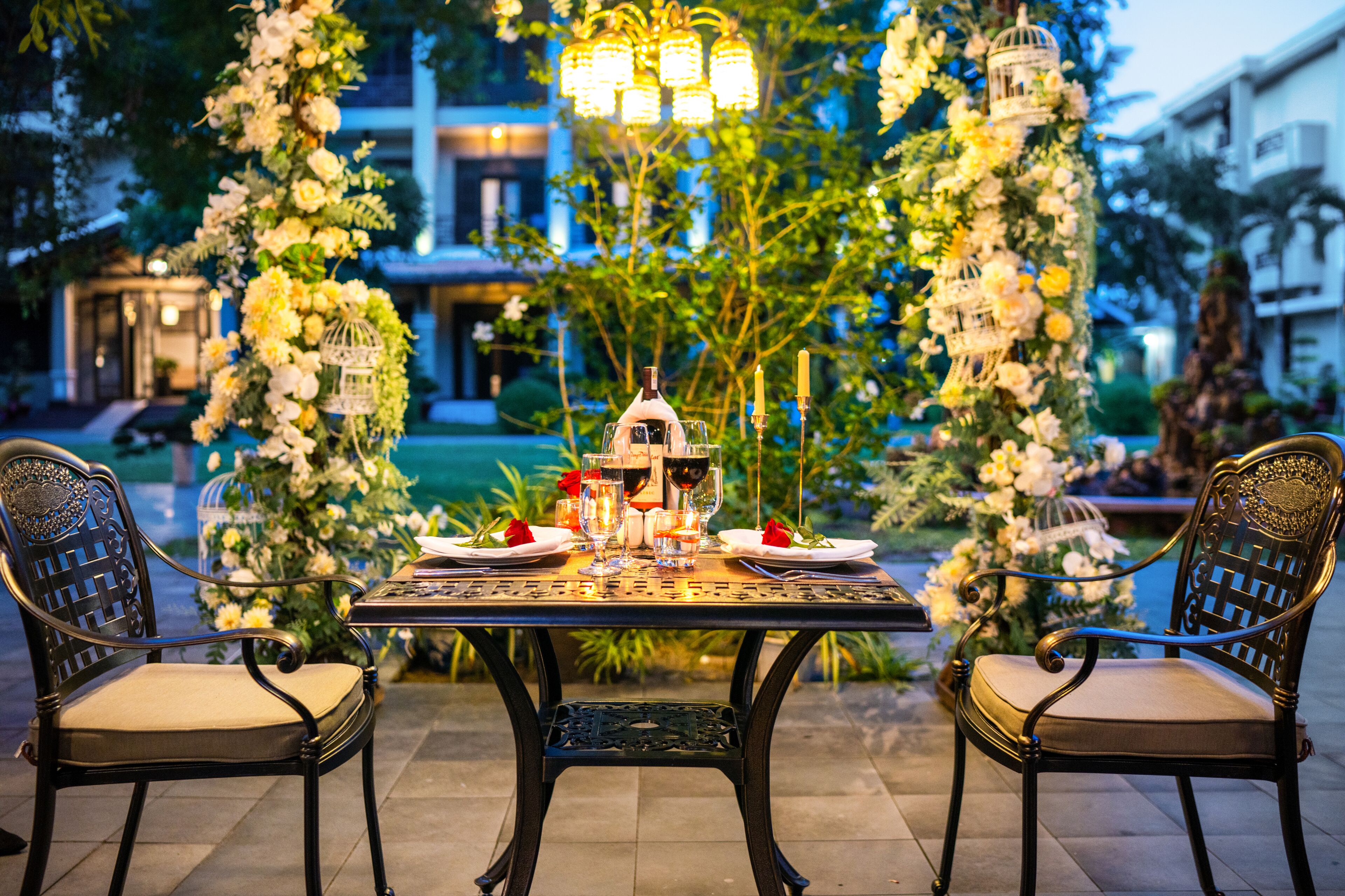 Un espace de restauration en plein air avec une table dressée pour deux personnes, entourée de chaises décoratives et d’arrangements floraux.